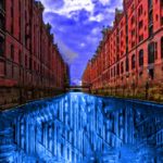 A.P. ASTRA - Cross: A canal flanked by tall red-brick buildings reflects a distorted, abstract blue pattern in the water, under a vivid, partly cloudy sky.