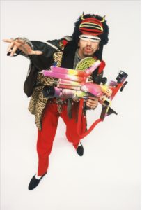 A person dressed in colorful, eclectic clothing and accessories—including red pants, a leopard print coat, and a black furry hat with horns—strikes a dramatic pose with a toy-like contraption, channeling the playful spirit of GEORGE DUBOSE - B-52s photography.