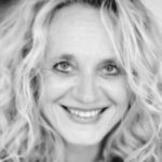A black and white portrait of Jana Dettmer with curly hair and a joyful smile, looking directly at the camera. Her eyes are expressive and she radiates happiness.