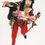 A person dressed in colorful, eclectic clothing and accessories—including red pants, a leopard print coat, and a black furry hat with horns—strikes a dramatic pose with a toy-like contraption, channeling the playful spirit of GEORGE DUBOSE - B-52s photography.