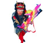 A man in colorful, eccentric clothing and sunglasses, reminiscent of a GEORGE DUBOSE - B-52s photoshoot, holds a large, brightly painted toy-like gun while standing against a white background.