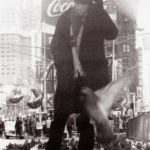 A person in a dark coat and hat stands among pigeons in a busy Times Square, with blurred birds flying nearby and a large Coca-Cola billboard behind—capturing the dynamic street style seen in GEORGE DUBOSE - B-52s photography.