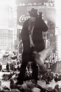 A person in a dark coat and hat stands among pigeons in a busy Times Square, with blurred birds flying nearby and a large Coca-Cola billboard behind—capturing the dynamic street style seen in GEORGE DUBOSE - B-52s photography.