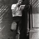 A man in a white t-shirt and dark pants leans against a wall, smoking a cigarette. His hat hangs on a pipe as he gazes at the camera in a shadowy, urban setting—an image reminiscent of GEORGE DUBOSE - B-52s photography.