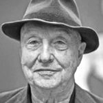 An older man with a slight smile wears a fedora hat and a blazer. The black-and-white photo, reminiscent of portraits of Georg Baselitz, highlights his facial wrinkles and calm expression against a blurred background.