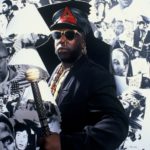 A man in dark clothing, sunglasses, and a black leather hat stands confidently holding a staff, posed in front of a wall covered with black-and-white photographs taken by GEORGE DUBOSE—legendary photographer for the B-52s and other icons.