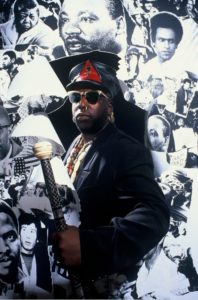 A man in dark clothing, sunglasses, and a black leather hat stands confidently holding a staff, posed in front of a wall covered with black-and-white photographs taken by GEORGE DUBOSE—legendary photographer for the B-52s and other icons.