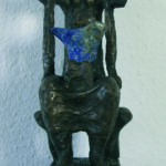 A bronze sculpture by Stephan Balkenhol, reminiscent of Atlas, depicts a seated human figure with raised arms and a blue stone embedded in the torso, all mounted on a reddish-brown rectangular base against a light textured wall.