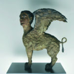 A sculpture inspired by STEPHAN BALKENHOL ATLAS features a mythological creature with a human head, lion’s body, and large wings, standing on a rectangular black base against a plain background.