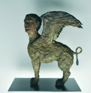 A sculpture inspired by STEPHAN BALKENHOL ATLAS features a mythological creature with a human head, lion’s body, and large wings, standing on a rectangular black base against a plain background.
