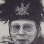 Eine Person, die eine Brille und einen großen, gefiederten Hut trägt, lächelt leicht, während sie das Hutband mit den Händen zurechtrückt. Das schwarz-weiße Bild erinnert an den spielerischen Geist des deutschen Malers Sigmar Polke in der zeitgenössischen Kunst.