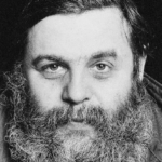 Schwarz-Weiß-Porträt eines Mannes mit dichtem dunklem Haar und buschigem Vollbart, der eine Jacke trägt und mit neutralem Blick direkt in die Kamera schaut.