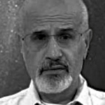 Schwarz-Weiß-Foto von Angelos Panagiotou, einem älteren Mann mit Glatze, grauem Bart und Schnurrbart, der eine Brille und ein helles Hemd mit Kragen trägt und direkt in die Kamera schaut.