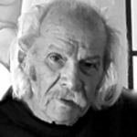 Ein älterer Mann mit weißem Haar und Schnurrbart, möglicherweise Christos Karas, schaut mit ernster Miene in die Kamera. Das Bild ist in Schwarz-Weiß gehalten und der Hintergrund ist unscharf.