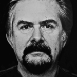 Schwarz-Weiß-Porträt eines Mannes mittleren Alters mit kurzem, zurückgekämmtem Haar, Schnurrbart und dunklem Ziegenbart, der mit einem ernsten Ausdruck, der an Fernando Boteros unverwechselbaren Stil erinnert, direkt in die Kamera blickt.