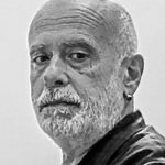 Schwarz-Weiß-Porträt des italienischen Malers Francesco Clemente, eines älteren Mannes mit Glatze, weißem Bart, Schnurrbart, dunkler Jacke und kleinem Ohrring, der vor einem schlichten Hintergrund ernst in die Kamera schaut.