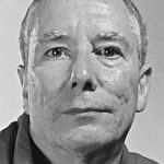 Graustufen-Nahaufnahme eines älteren Mannes mit kurzen Haaren, der ein Hemd mit Kragen trägt und mit neutralem Blick direkt in die Kamera schaut.