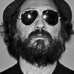 Eine Person mit einem dichten Bart, die wie Mr. Brainwash aussieht, trägt eine Piloten-Sonnenbrille, einen Hut und ein dunkles Hemd und schaut direkt in die Kamera. Das Bild ist in schwarz-weiß gehalten.