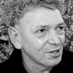 Ein Schwarz-Weiß-Foto zeigt Robert Combas mit lockigem Haar, der leicht zur Seite schaut und lächelt. Im Hintergrund ist ein mosaikartiges Muster zu sehen.