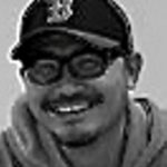 Eine Person, möglicherweise Seung-Koo Lee, die eine Brille, einen Schnurrbart, eine Baseballmütze mit dem Buchstaben "B" und einen Kapuzenpulli trägt, lächelt auf diesem Schwarzweißbild in die Kamera.