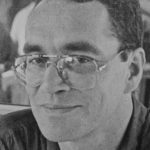 Ein Schwarz-Weiß-Foto von Stephan Runge, einer Person mit kurzen dunklen Haaren, einer großen Brille und einem Hemd mit Kragen, die in die Kamera schaut und sanft lächelt.