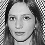 Schwarz-Weiß-Foto einer Frau mit langem glattem Haar, die Ohrringe trägt und direkt in die Kamera schaut. Im Hintergrund ist ein Muster aus gleichmäßig verteilten schwarzen Punkten zu sehen, das an den abstrakten Kunststil von Tauba Auerbach erinnert.