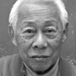 Ein älterer Mann mit weißem Haar und einem neutralen Gesichtsausdruck blickt direkt in die Kamera. Er trägt ein Hemd mit Kragen und einen Pullover. Das Bild ist in schwarz-weiß gehalten.